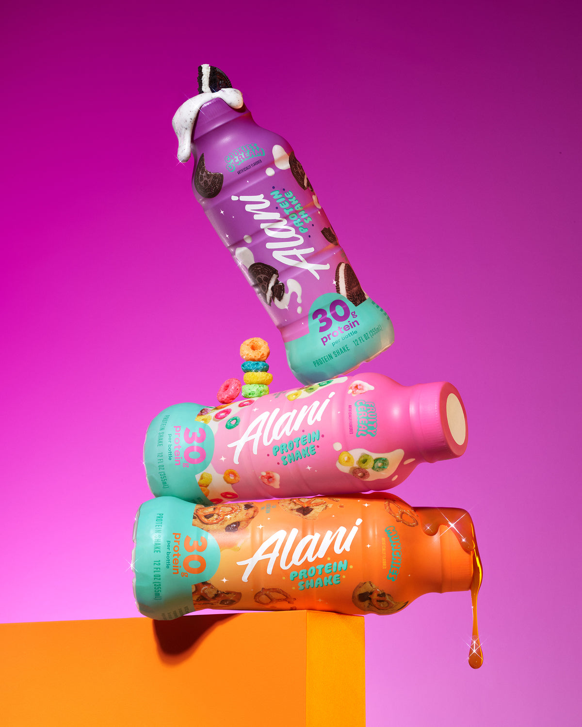 A trio of Alani Nu 30-gram protein shakes: Cookies &amp; Cream, Fruity Cereal, and Munchies, stacked on an orange ledge against a purple gradient background. 