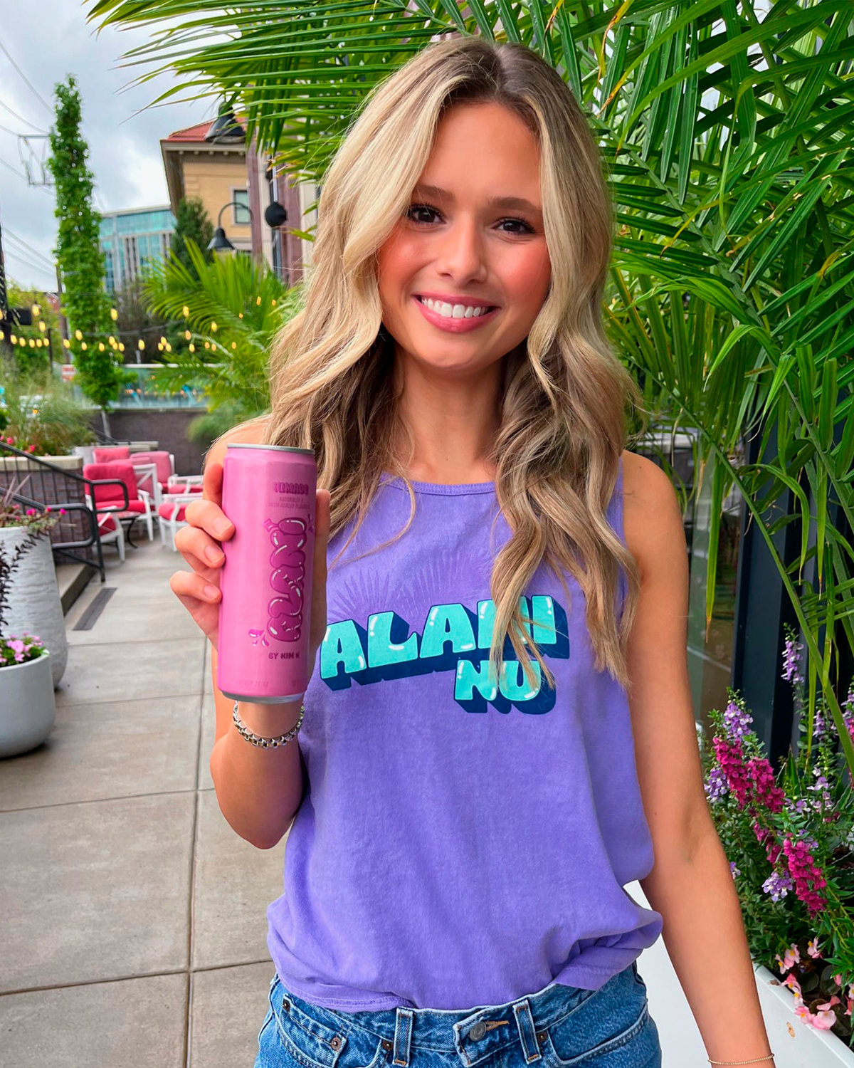 A woman with long blonde hair, wearing the limited-edition Purple Palm Tank from Alani Nu and blue jeans, holds a can of a pink beverage while standing outdoors near greenery and buildings.