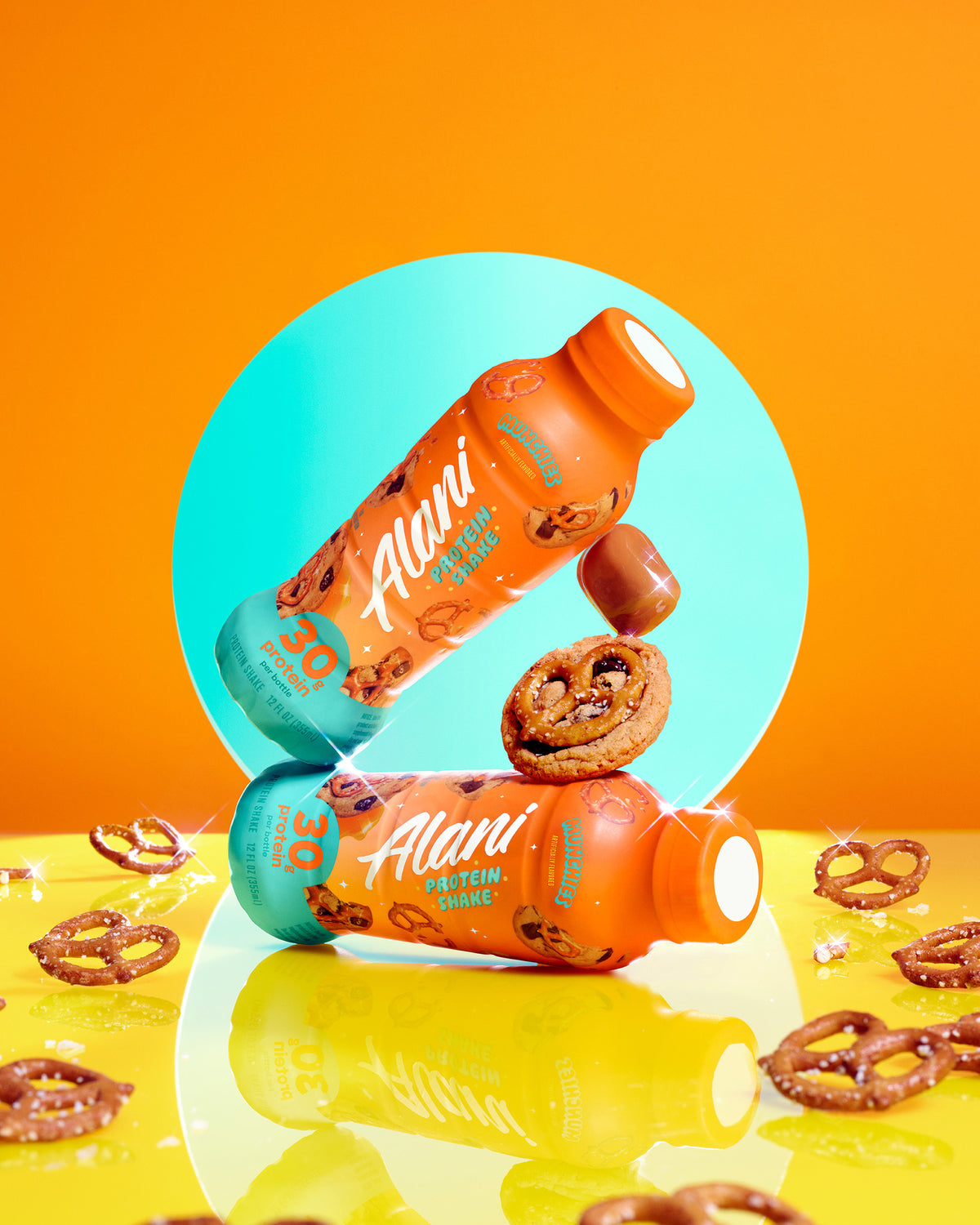 A whimsical image of two bottles of Munchies protein stacked on their sides and resting on a reflective yellow surface, surrounded by cookies, pretzels, and caramels.