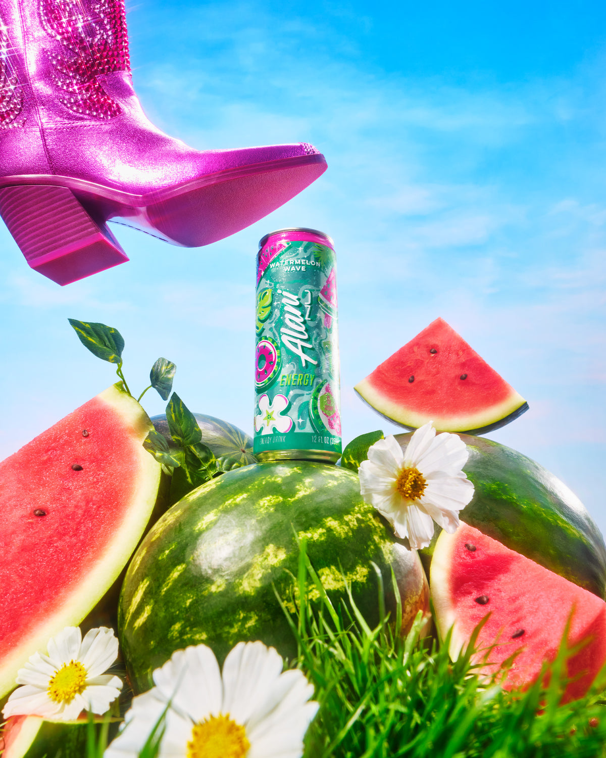 A can of Alani Nu Watermelon Wave Energy sitting atop a whole watermelon in a plot of grass surrounded by sliced watermelon, flowers, and vines. A pink cowboy boot is just above the can as if it’s going to smash it.  