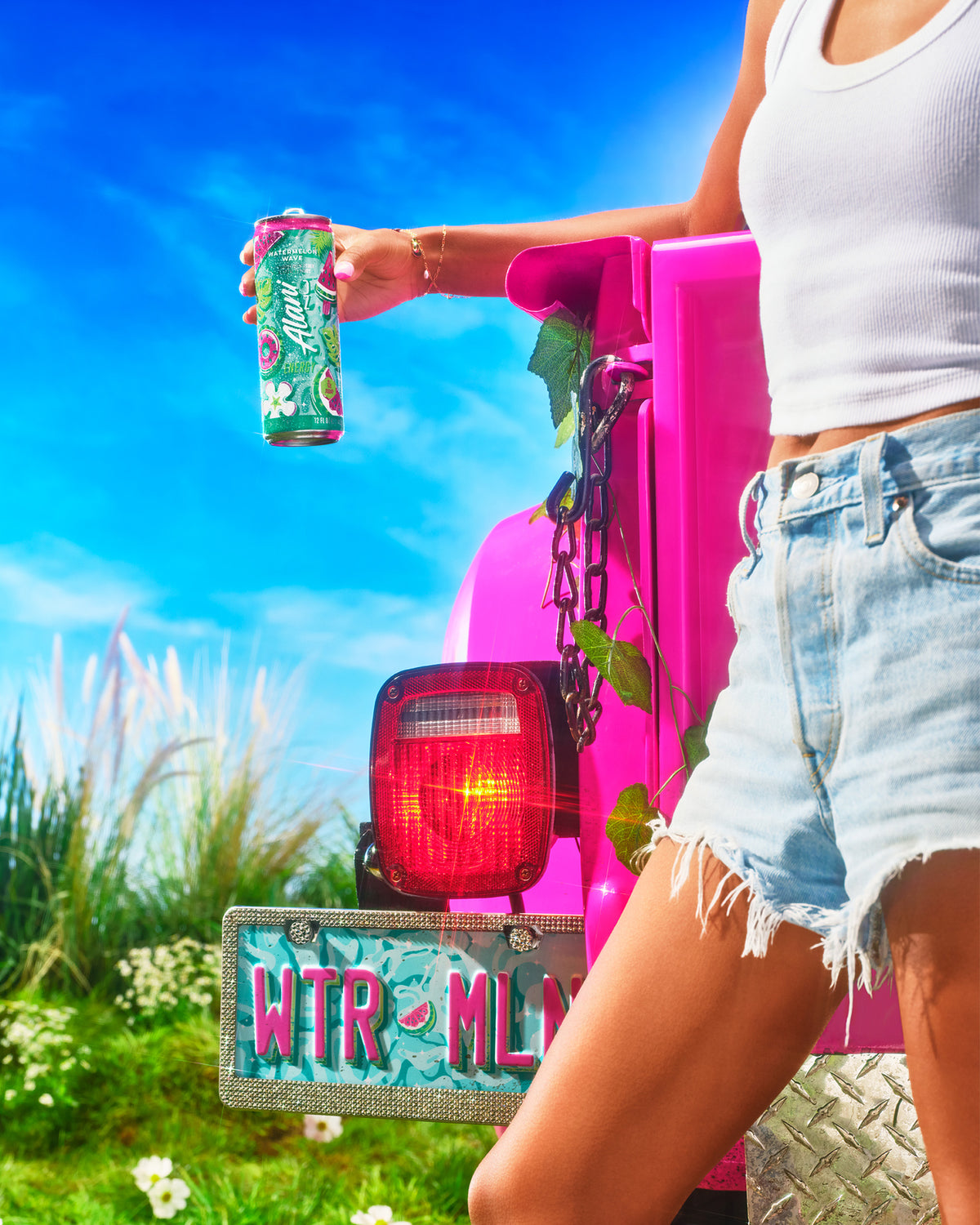 A woman shown from chest to mid-shin standing in front of a pink vintage truck holding a can of Alani Nu Watermelon Wave Energy. She’s wearing hot pink iridescent cowboy boots and the license plate reads WTR-MLN.  